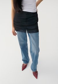 Falda negra transparente sobre vaqueros rectos de color azul claro, combinada con zapatos de punta roja y hebillas. Textura suave y diseño en capas.