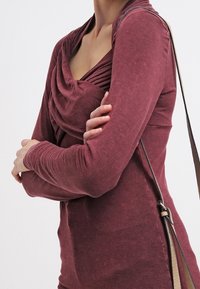 Bordeaux långärmad topp med draperad halsringning och texturerade ärmar. Bäraren har armarna korsade och en axelväska med sned rem.