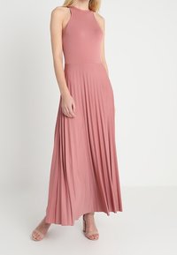 Sukienka maxi z miękkiego różowego materiału, z dopasowanym górnym partem i rozkloszowanym plisowanym dołem. Gładka faktura, bez rękawów, sięgająca do podłogi.