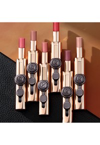 Seks leppestifter med gullfargede metallkapsler, svarte knapper og varierende nyanser av rosa og nude. De er arrangert på en teksturert svart overflate.