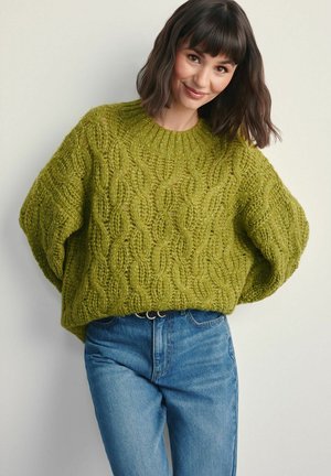 Zielony sweter z dzianiny z wzorem warkocza i oversize'owymi rękawami, zestawiony z niebieskimi dżinsami. Teksturowana tkanina i prążkowane detale przy dekolcie.