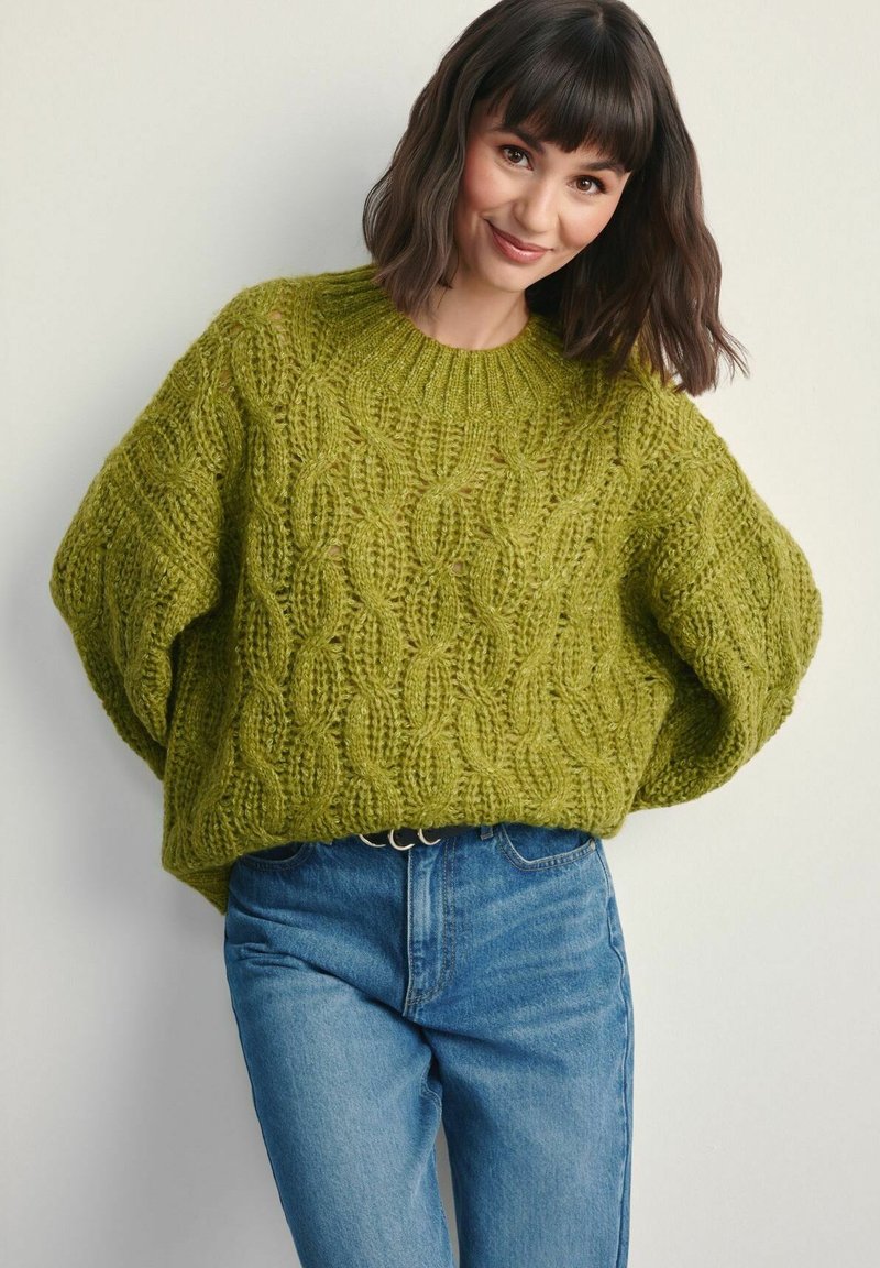 Zielony sweter z dzianiny z wzorem warkocza i oversize'owymi rękawami, zestawiony z niebieskimi dżinsami. Teksturowana tkanina i prążkowane detale przy dekolcie.