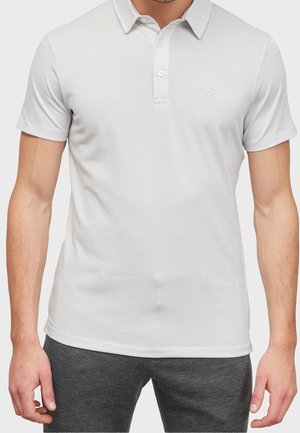 Mann trägt ein weißes Poloshirt mit kurzen Ärmeln, Kragen und zwei Knöpfen, kombiniert mit dunkelgrauer Hose.