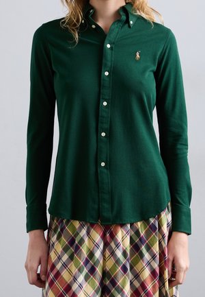 Femme portant une chemise boutonnée vert foncé avec un petit logo brodé et une jupe à carreaux multicolore, debout devant un fond uni.
