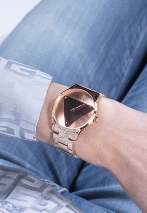 Ceas de mână cu brățară metalică aur roz și față neagră triunghiulară care afișează ora pe încheietura mâinii stângi a unei persoane, îmbrăcată în blugi albaștri și cămașă cu model.