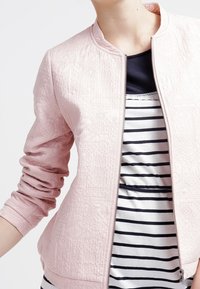 Chaqueta bomber acolchada de color rosa claro con cuello redondo y cierre de cremallera. Debajo, se puede ver una camiseta a rayas en colores azul marino y blanco.