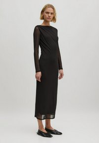 Schwarzes langärmliges Kleid aus glattem Stoff mit drapiertem Design. Transparente Ärmelakzente und ein fließender Rock bis zur Mitte der Wade. Kombiniert mit flachen Schuhen.