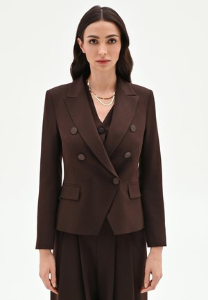 Donna con capelli castano scuro indossa un blazer doppiopetto marrone scuro su misura e pantaloni coordinati, accessoriati con collane a catena d'oro.