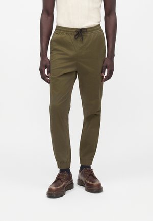 Homme debout portant un pantalon de jogging vert olive avec cordon et des chaussures marron à lacets avec des chaussettes noires, sur un fond blanc uni.