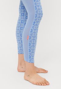 Pernas inferiores e pés a usar leggings azul estampadas com uma pequena etiqueta vermelha, mostrando duas pequenas tatuagens nos tornozelos, de pé sobre uma superfície branca.