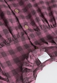 Combinaison bébé à carreaux roses et noirs, avec un col à volants, une fermeture à boutons et une texture douce. Étiquette visible avec le nom de la marque "NEXXT".
