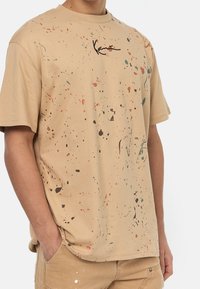 Béžové bavlněné tričko s volným vzorem spláchnuté barvy v odstínech modré, oranžové a černé. Černé logo potištěné na přední straně. Krátké rukávy.