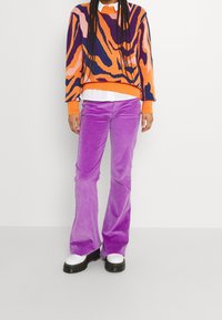 Suéter de gran tamaño y colorido con patrones abstractos en naranja, morado y rosa, combinado con pantalones de pana morados de pierna acampanada y zapatos de suela gruesa negros.