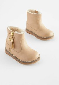 Botas de tobillo de cuero de color beige claro con forro de pelo sintético, detalles en hardware dorado, cremallera lateral y suela de goma marrón con detalles de costura blanca.