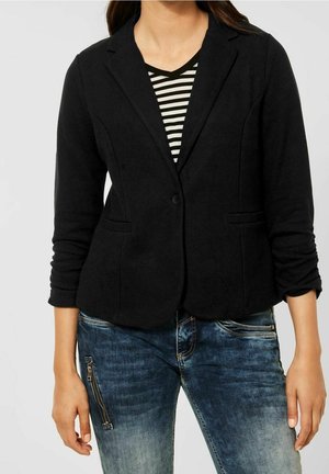 Osoba ubrana w czarną marynarkę na jeden guzik na czarno-białą, paskowaną koszulkę z dekoltem w serek oraz niebieskie, wyblakłe dżinsy z kieszenią na zamek.