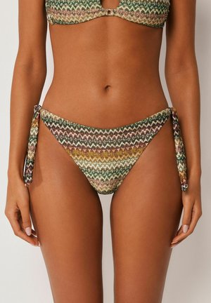 Kobieta ubrana w bikini w wielokolorowy wzór zygzaków, z wiązanymi bokami dołu i pasującym stanikiem, widziana od talii do górnej części ud.