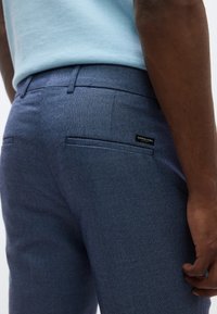 Ανοιχτό μπλε τοπ φορεμένο με υφασμάτινο σκούρο μπλε παντελόνι με δύο πίσω τσέπες και μικρή μαύρη ετικέτα στην δεξιά τσέπη.