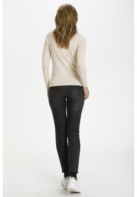 Beige top med långa ärmar i ljus nyans kombinerad med mörkgrå tajta jeans, med en åtsittande silhuett och vita sneakers, sedd bakifrån.