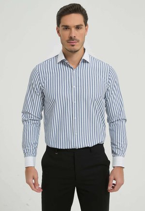 Homme portant une chemise blanche à manches longues avec des rayures verticales bleues et un pantalon noir, debout devant un fond clair uni.