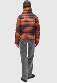Pohodlná puffer bunda s vícebarevným gradientem v oranžové, hnědé a černé barvě. Má vysoký límec a prošívaný design. Nosí se s šedými kordurovými kalhotami.