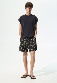Čierne tielko bez rukávov, vzorované čierne šortky so zlatými slnečnými motívmi a čierne šľapky. Neformálny letný outfit.
