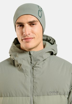 Homme portant une veste à capuche à carreaux vert clair, fermée par une fermeture éclair, et un bonnet assorti avec un logo sur le côté et sur la poitrine.