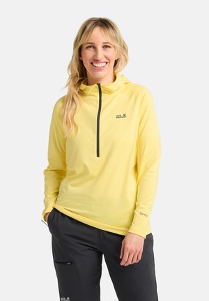 Kurzärmlige, kapuzenlose, gelbe Jacke mit einem Viertel-Reißverschluss, ausgestattet mit einem schwarzen Logo und strukturiertem Stoff, kombiniert mit dunklen Hosen.