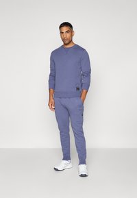 Sweatshirt lavande clair avec des poignets et une taille côtelés, associé à un pantalon de survêtement assorti avec des poches latérales et un détail de marque. Porteur en baskets blanches.