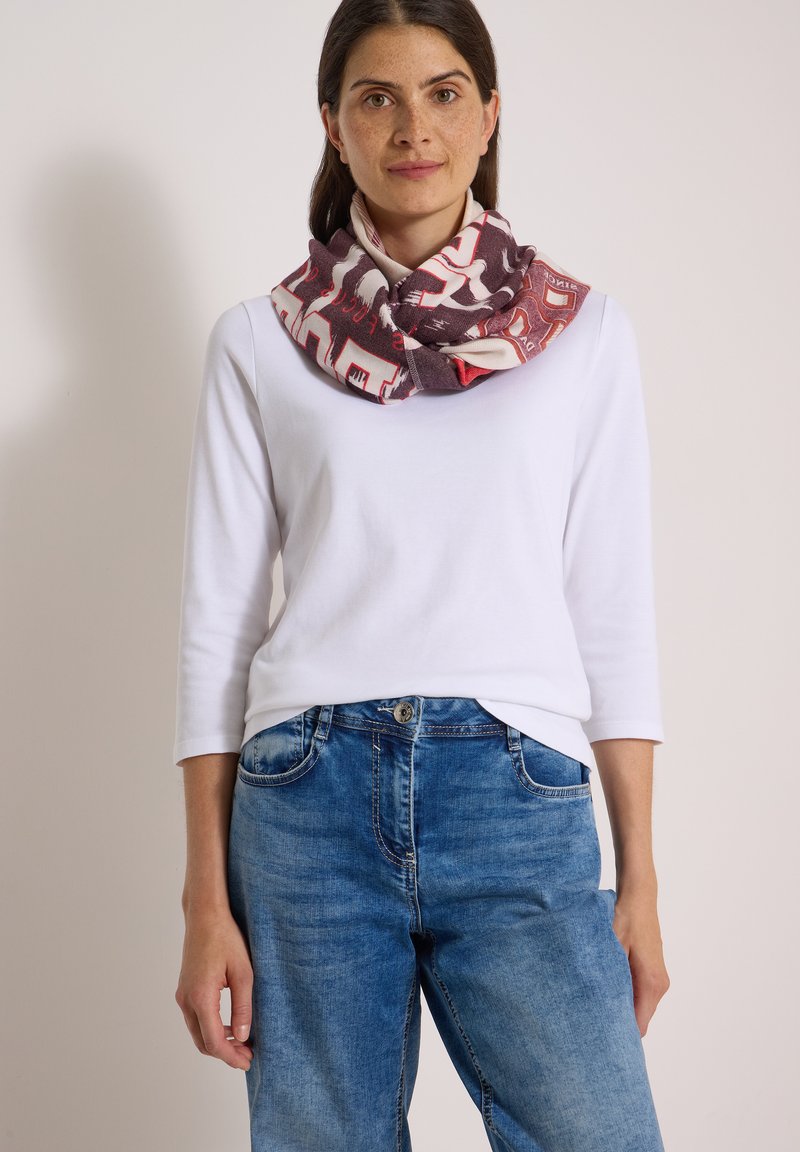 Femme portant un haut blanc à manches longues, un jean bleu et une écharpe multicolore à motifs autour du cou, debout devant un fond uni.
