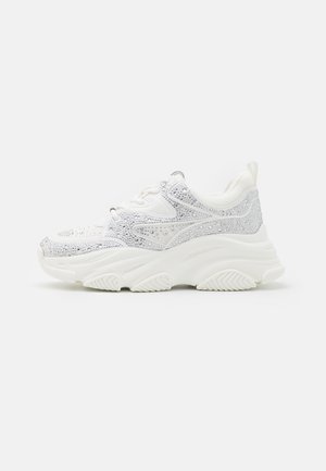 Biały masywny sneaker z grubą podeszwą, pokryty srebrzystymi kryształkami i teksturowanym bieżnikiem na jednolitym tle.