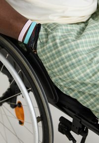 Groen en crèmekleurige geblokte stof bedekt de benen van een persoon in een rolstoel. Meerdere armbanden worden om de pols gedragen, naast een zwarte armleuning van de rolstoel.