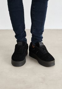 Baskets en suède noir avec une semelle en caoutchouc épaisse, présentant des bouts ronds, des lacets plats, et une petite étiquette logo sur la languette. Portées avec un jean foncé.