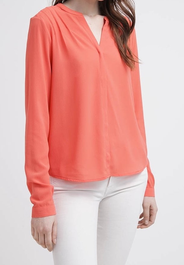 Blouse corail à manches longues en tissu léger, avec un décolleté en V et de subtils plis à l'avant, assortie à un pantalon blanc.
