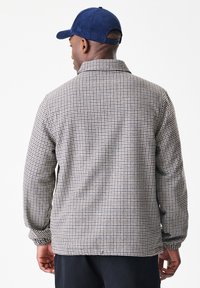 Άντρας φοράει μπλε καπέλο και μπουφάν με μακρύ μανίκι με καφέ και άσπρη καρό σχεδία, στέκεται με την πλάτη του στραμμένη προς την κάμερα.