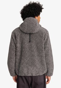 Pull en polaire gris avec un col haut et une capuche. Il présente une texture douce, une coupe décontractée et un détail en cordon noir à l'arrière.