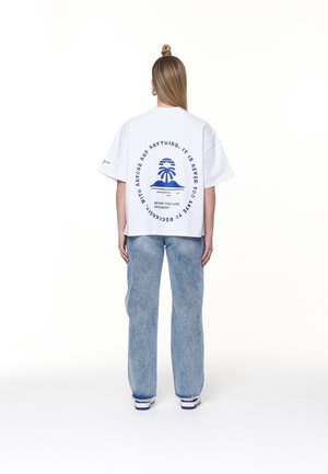 Osoba z długimi włosami związanymi w kok nosi białą, oversize'ową koszulkę z niebieską grafiką palmy i tekstem, w połączeniu z jasnoniebieskimi dżinsami i trampkami.
