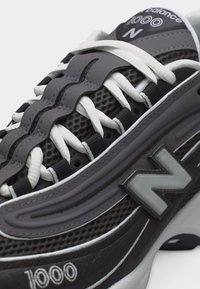 Gros plan sur une basket New Balance 1000 gris foncé avec des lacets blancs, une tige en mesh texturé et le logo "N" de la marque sur le côté.