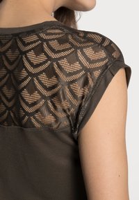 Top de color marrón oscuro que presenta un escote de encaje transparente con un patrón geométrico en forma de concha. Mangas cortas con tela suave debajo del escote.