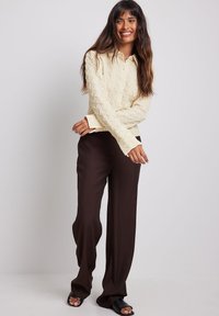 Camicetta a maniche lunghe di colore crema con colletto, abbinata a pantaloni larghi marrone scuro e calzature nere con punta aperta.