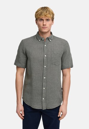 Junger Mann mit kurzem blondem Haar, der ein strukturiertes graues Kurzarmhemd mit Knöpfen und dunkle Hose trägt, steht vor einem weißen Hintergrund.