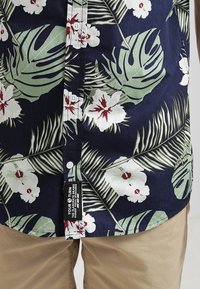Krátký rukáv košile z lehké látky, s tmavě modrým pozadím, velkými bílými květy, zelenými listy a malými bílými knoflíky.