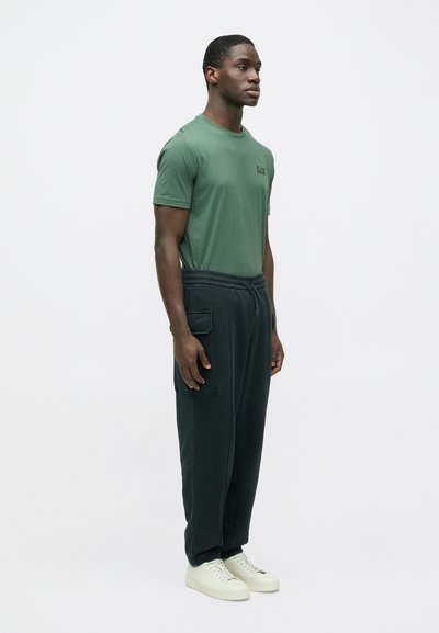 Zelené tričko s krátkym rukávom a logom na hrudníku, skombinované s voľnými čiernymi teplákmi s bočnými vreckami a bielymi teniskami.