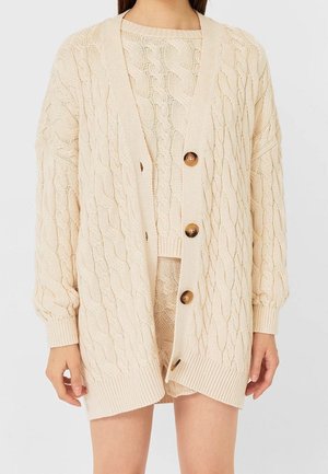 Woman wearing beige cable knit cardigan with large buttons over matching knit shorts and sweater against pale background.