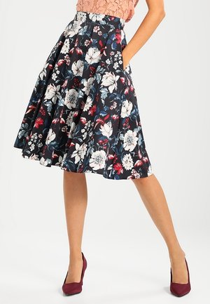 Femme portant une jupe plissée noire mi-longue ornée d'un imprimé floral blanc, rouge et bleu, associée à des escarpins bordeaux.