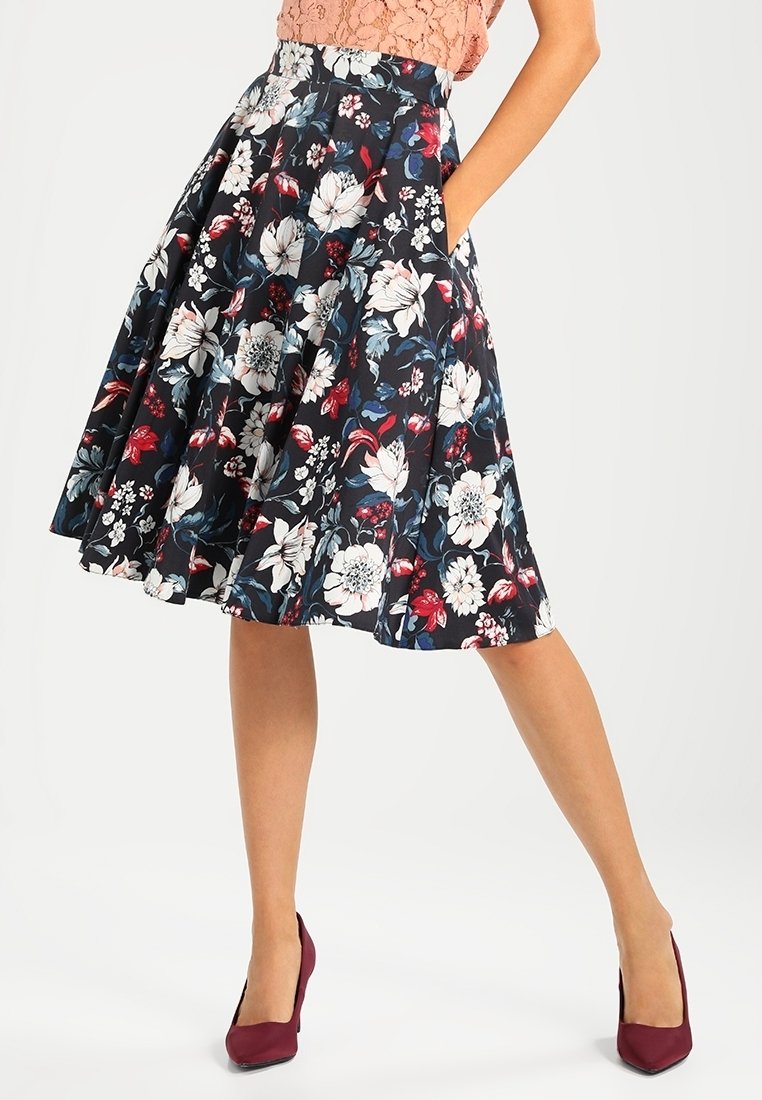 Femme portant une jupe plissée noire mi-longue ornée d'un imprimé floral blanc, rouge et bleu, associée à des escarpins bordeaux.