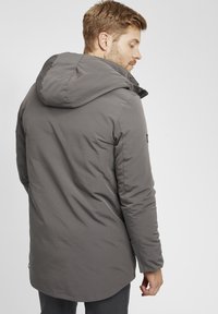 Chaqueta acolchada gris con capucha, que presenta una textura suave, mangas largas y un dobladillo ligeramente curvado. Sin detalles de hardware visibles.