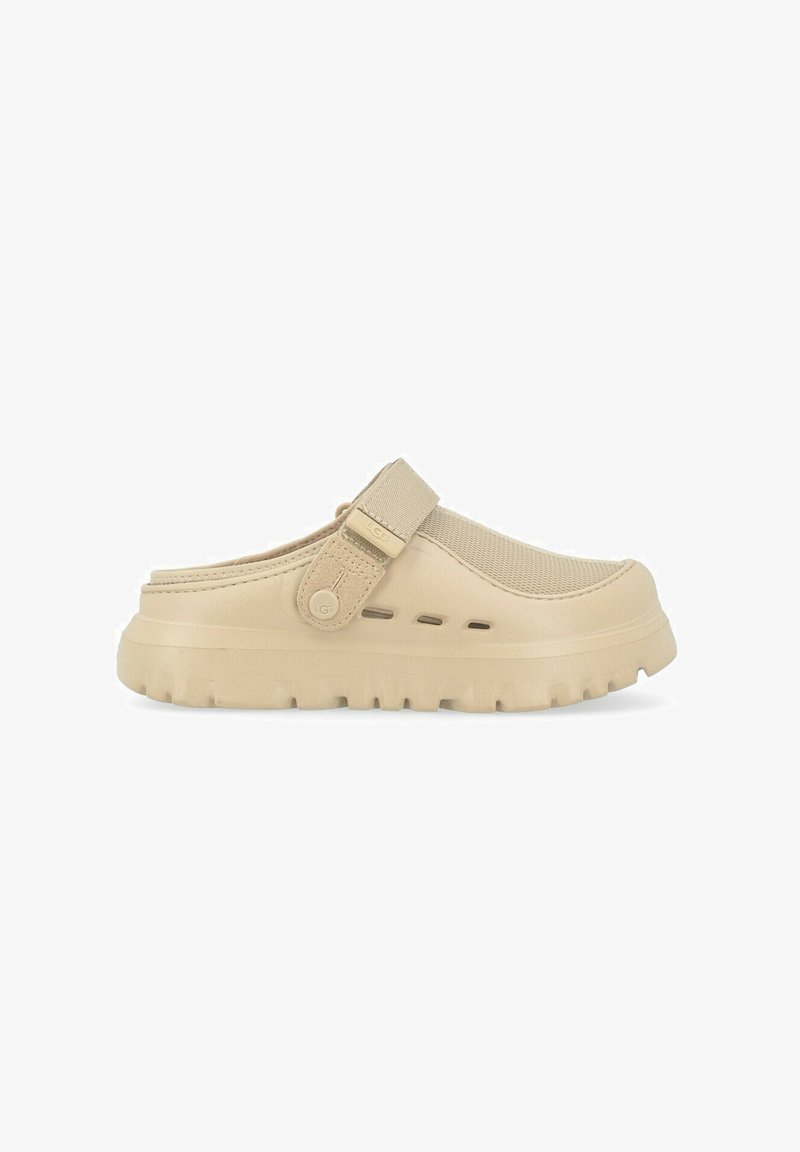 Zapatilla tipo zueco color beige con parte superior texturizada, correa ajustable, suela gruesa y orificios de ventilación a lo largo del costado. Material ligero.
