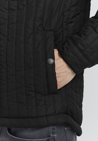 Giacca nera trapuntata caratterizzata da un design testurizzato e imbottito, tasca laterale con chiusura a scatto e una finitura in tessuto liscia.