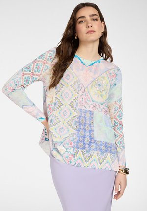 Donna con capelli lunghi castani che indossa una blusa pastello multicolore a fantasia e una gonna viola chiaro, accessoriata con braccialetti e orecchini dorati.
