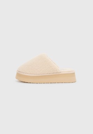 Beige plush slipper with a rounded upper, soft texture, and a thick tan platform sole featuring a textured bottom for grip.